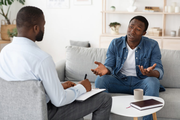 A stressed man engages in conversation with another man while seated on a couch, highlighting their serious therapy discussion.
