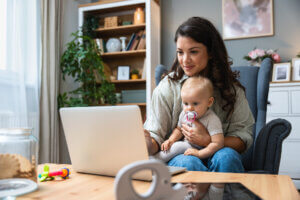 Young mother working on a laptop while holding her baby, symbolising the challenges and self-doubt of balancing work and motherhood